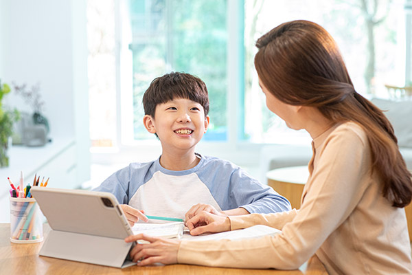 태블릿이 놓여진 책상에 앉은 여성과 남자아이가 눈을 맞추고 있는 모습 