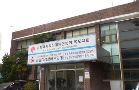 목포시각장애인생활이동지원센터 건물 측면으로 한국시각장애인연합회 목포지회 부설 목포시 시각장애인 생활이동지원센터 라고 명시된 간판과 (사)한국여성장애인 전남지부 전남여성장애인연대 부설 목포여성장애인성폭력상담소 부설 전남여성장애인 어울림센터 라고 명시된 간판이 함께 걸려있는 모습