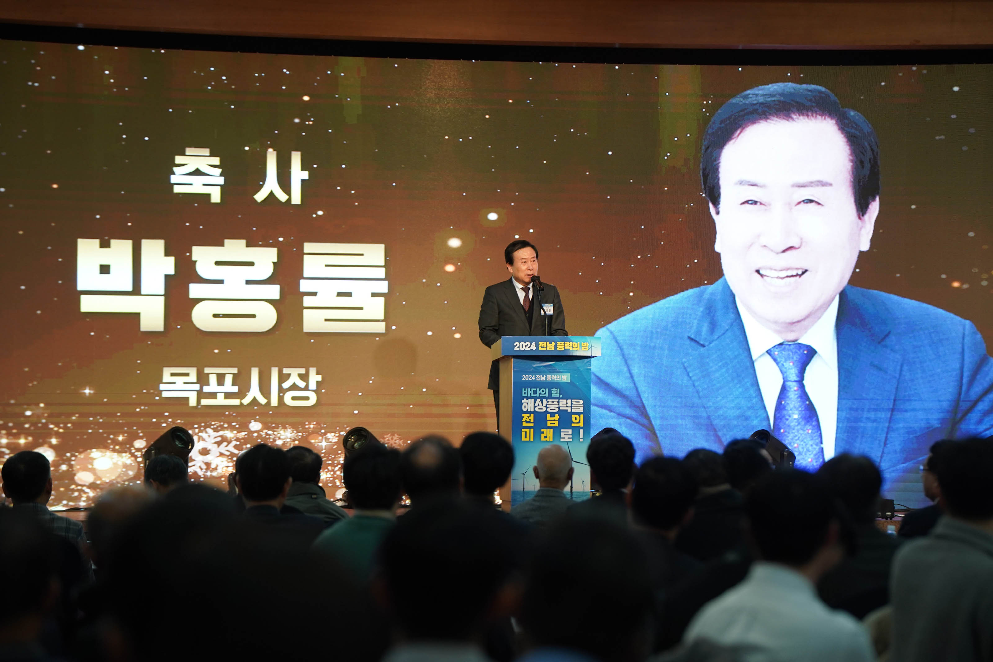 '축사 박홍률 목포시장'이라는 문구가 적힌 대형 스크린 앞 단상에서 박홍률 시장이 마이크 앞에 서서 연설하고 있는 모습. 단상 앞에는 '2024 전남 풍력의 밤'이라는 행사명이 적혀 있고, 무대 아래에는 청중들이 빼곡히 앉아 있는 모습이 보인다.