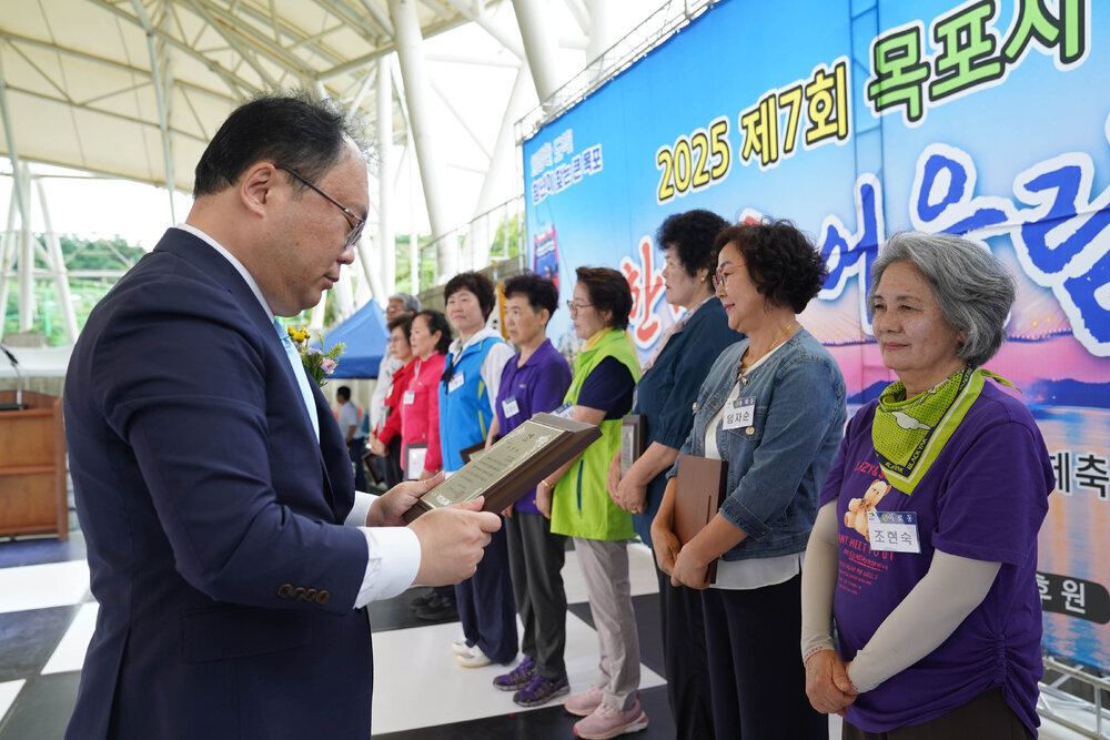 정장을 입고 안경을 낀 남성이 흰색 팔토시를 끼고 보라색 반팔티를 입은 조현숙에게 패를 수여하고 있다