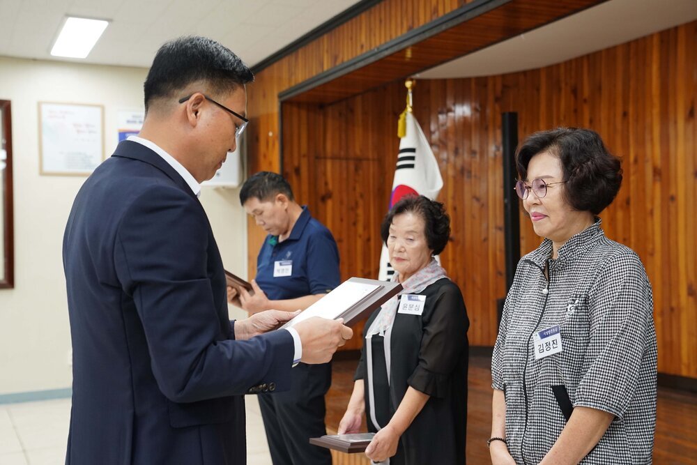 정장 차림의 관계자가 체크무늬 상의를 입은 여성 수상자에게 패를 전달하며 읽고 있는 모습이다.