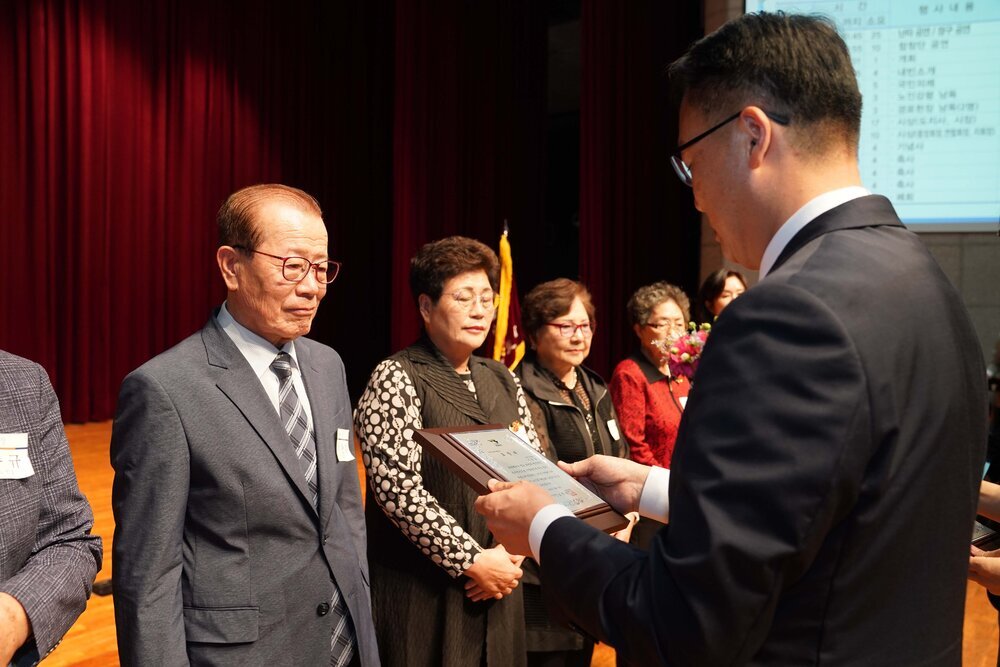 정장 차림의 관계자가 정장을 입은 남성 수상자에게 패를 전달하며 읽고 있는 모습이다.