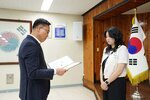 정장 차림의 관계자가 아이보리색 상의를 입은 여성 수상자에게 패를 전달하며 읽고 있는 모습이다.