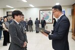 2026년 상반기 4급이하 정기인사로 정장을 입은 남성이 실내 행사장에서 한 남성에게 상장을 전달하며 내용을 읽고 있고, 남성은 안경을 쓰고 회색 울 자켓에 넥타이를 맨 상태로 사원증을 착용하고 두 손을 모으고 서 있으며 뒤쪽에는 참석자들이 행사를 지켜보고 있다.