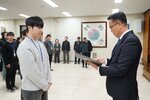 2026년 상반기 4급이하 정기인사로 정장을 입은 남성이 실내 행사장에서 한 남성에게 상장을 전달하며 내용을 읽고 있고, 남성은 밝은 회색 라운드 니트를 입고 사원증을 착용한 채 두 손을 모은 상태로 서 있으며 뒤쪽으로 참석자들이 줄지어 서서 행사를 지켜보고 있다.