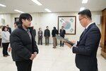 2026년 상반기 4급이하 정기인사로 정장을 입고 안경을 쓴 남성이 실내 행사장에서 한 남성에게 상장을 전달하며 내용을 읽고 있고, 남성은 검은 패딩을 입고 두 손을 모은 채 서 있으며 뒤쪽에는 참석자들이 줄지어 서서 행사를 지켜보고 있다.