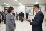 2026년 상반기 4급이하 정기인사로 정장을 입고 안경을 쓴 남성이 실내 행사장에서 한 여성에게 상장을 전달하며 내용을 읽고 있고, 여성은 회색 자켓을 입고 두 손을 모은 채 서 있으며 뒤쪽에는 여러 사람들이 줄지어 서서 행사를 지켜보고 있다.